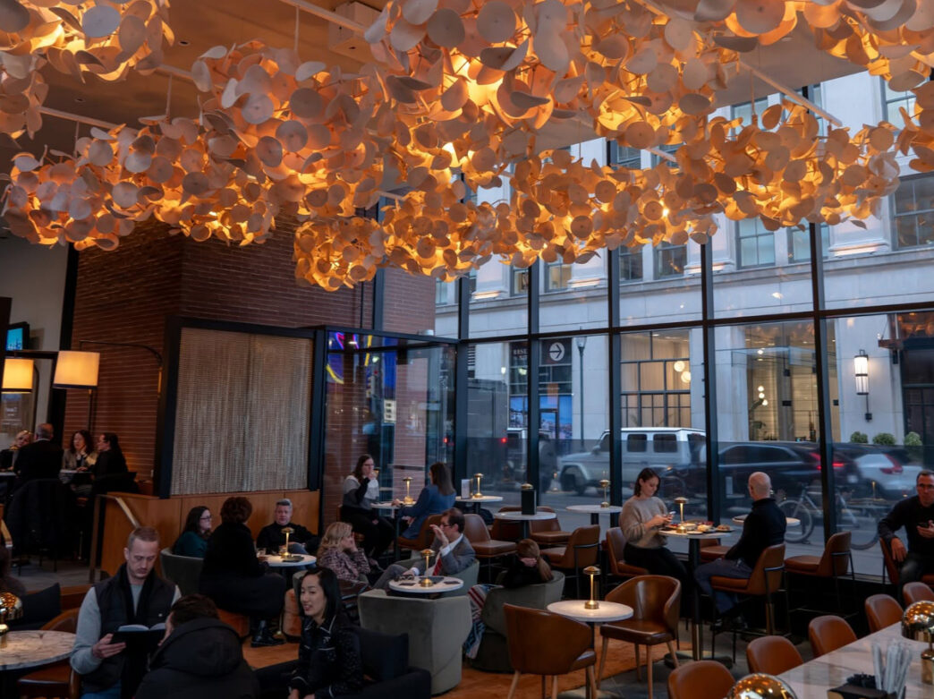 Diners gather at tables beneath a dramatic illumined ceiling installation in a modern dining room with floor to ceiling windows.