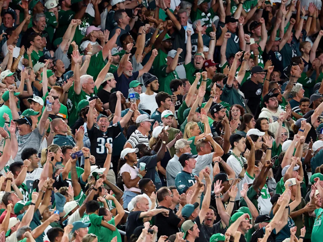 A packed crowd of Philadelphia Eagles fans dressed in green jerseys and team apparel cheers with raised arms and clapping hands during a game.