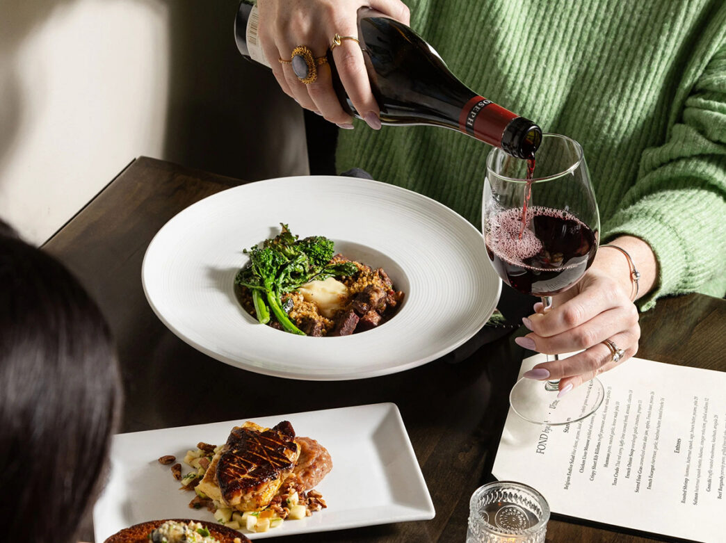 A diner wearing a green sweater pours red wine into a stemmed wine glass alongside artfully plated dishes and a dinner menu at Fond.
