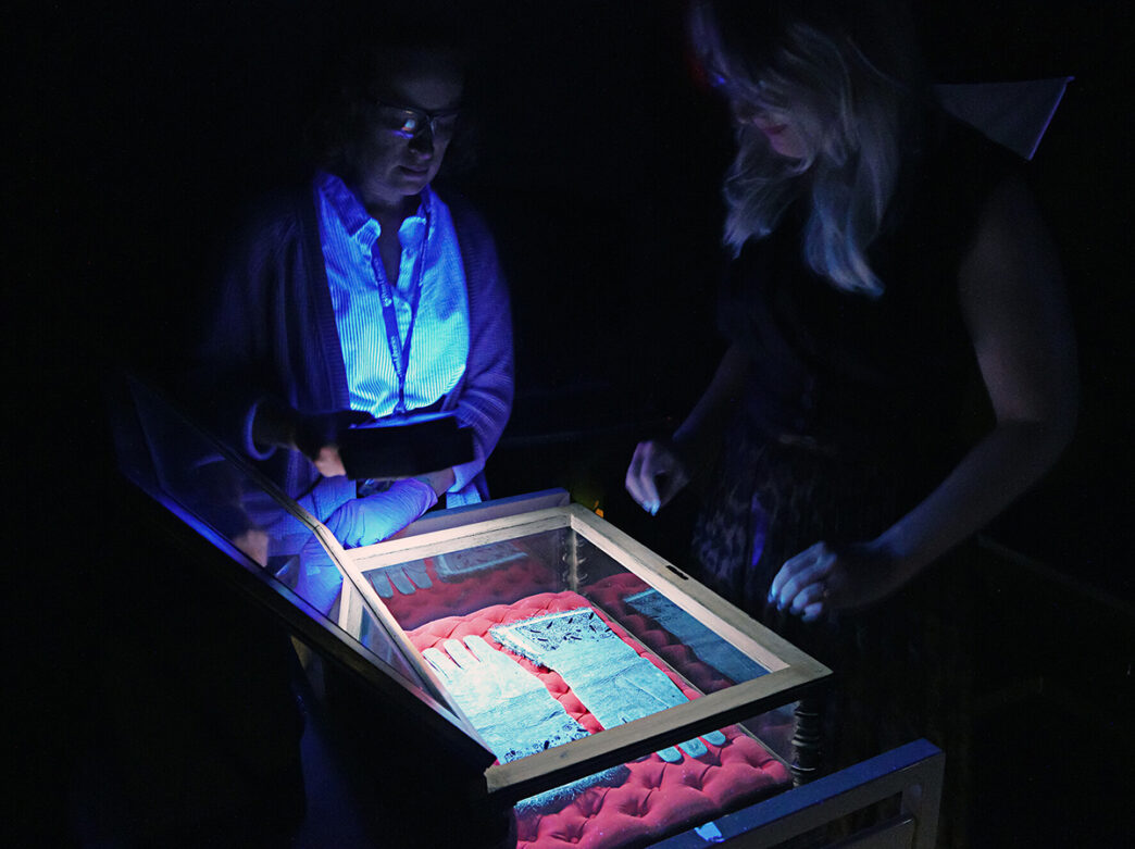 Two visitors examine a pair of historic gloves displayed in a glass case under dramatic ultraviolet lighting.