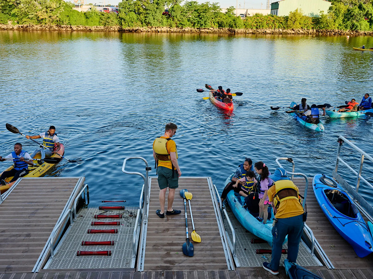 Visitors wearing life jackets and water shoes launch kayaks from a dock while other kayakers paddle on the river on a sunny day.