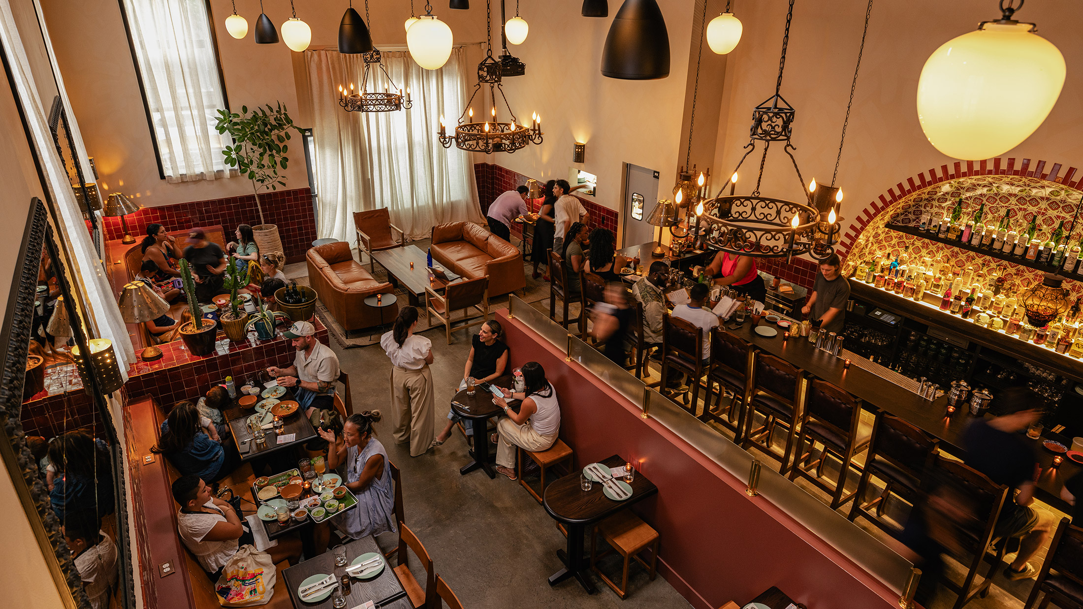 Diners fill the warm, colorful interior of Mixteca, with guests seated at tables and a long bar beneath ornate chandeliers and pendant lights.