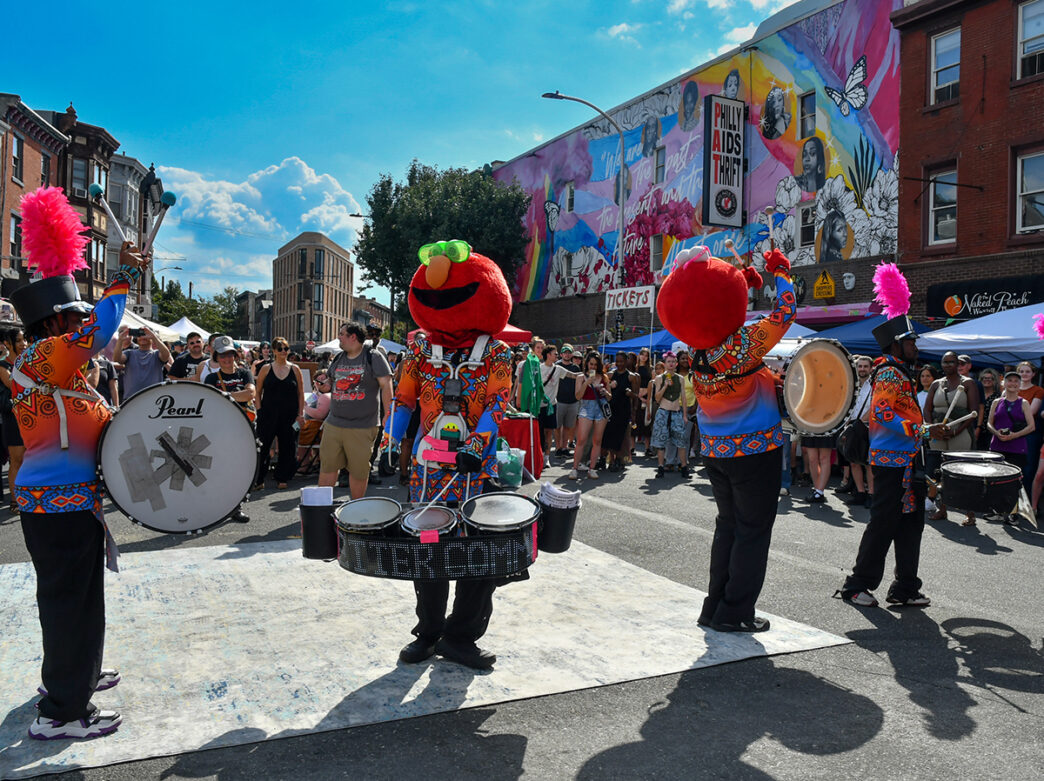 Performers in colorful uniforms and oversized Elmo character heads play drums for a crowd at the Philly AIDS Thrift Block Party, with a large mural visible in the background.