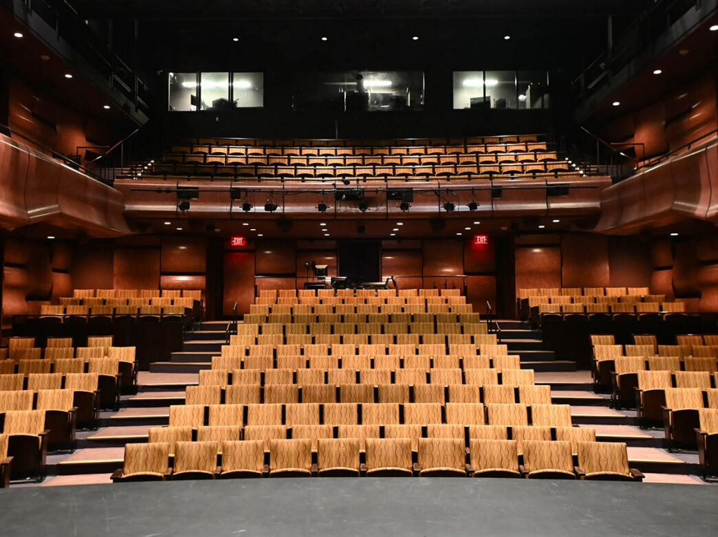 Un moderno auditorio de teatro con butacas de color beige dispuestas en gradas, un balcón superior y cálidas paredes revestidas de paneles de madera, visto desde el escenario.