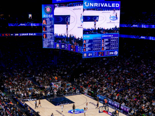 A packed arena surrounded a basketball court during an Unrivaled game between the Rose and Lunar Owls, with the center scoreboard displaying the live action and score.