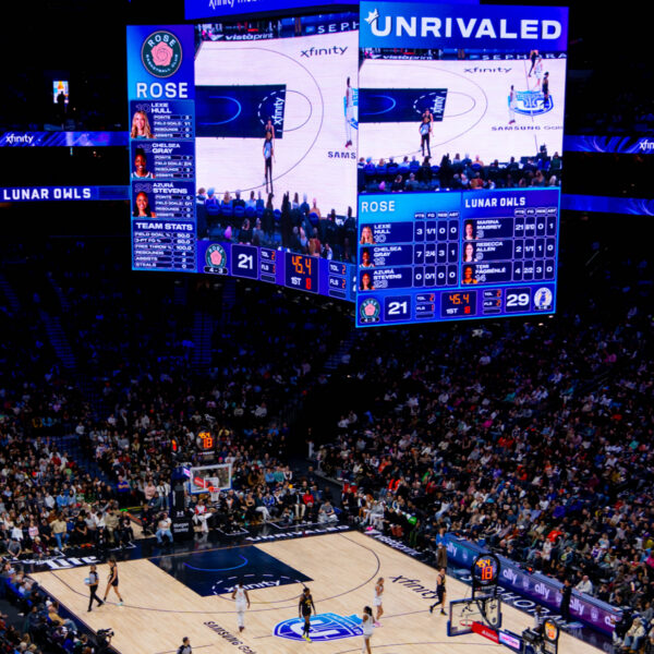 A packed arena surrounded a basketball court during an Unrivaled game between the Rose and Lunar Owls, with the center scoreboard displaying the live action and score.
