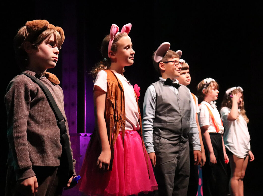 A group of children wearing animal ears and costumes stand in a row under stage lights during a youth theater performance.