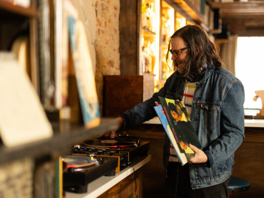 A meaning wearing a jean jacket and glasses plays a vinyl record on a turntable, while holding several record sleeves beside a shelf of vinyl records in a warmly lit bar.