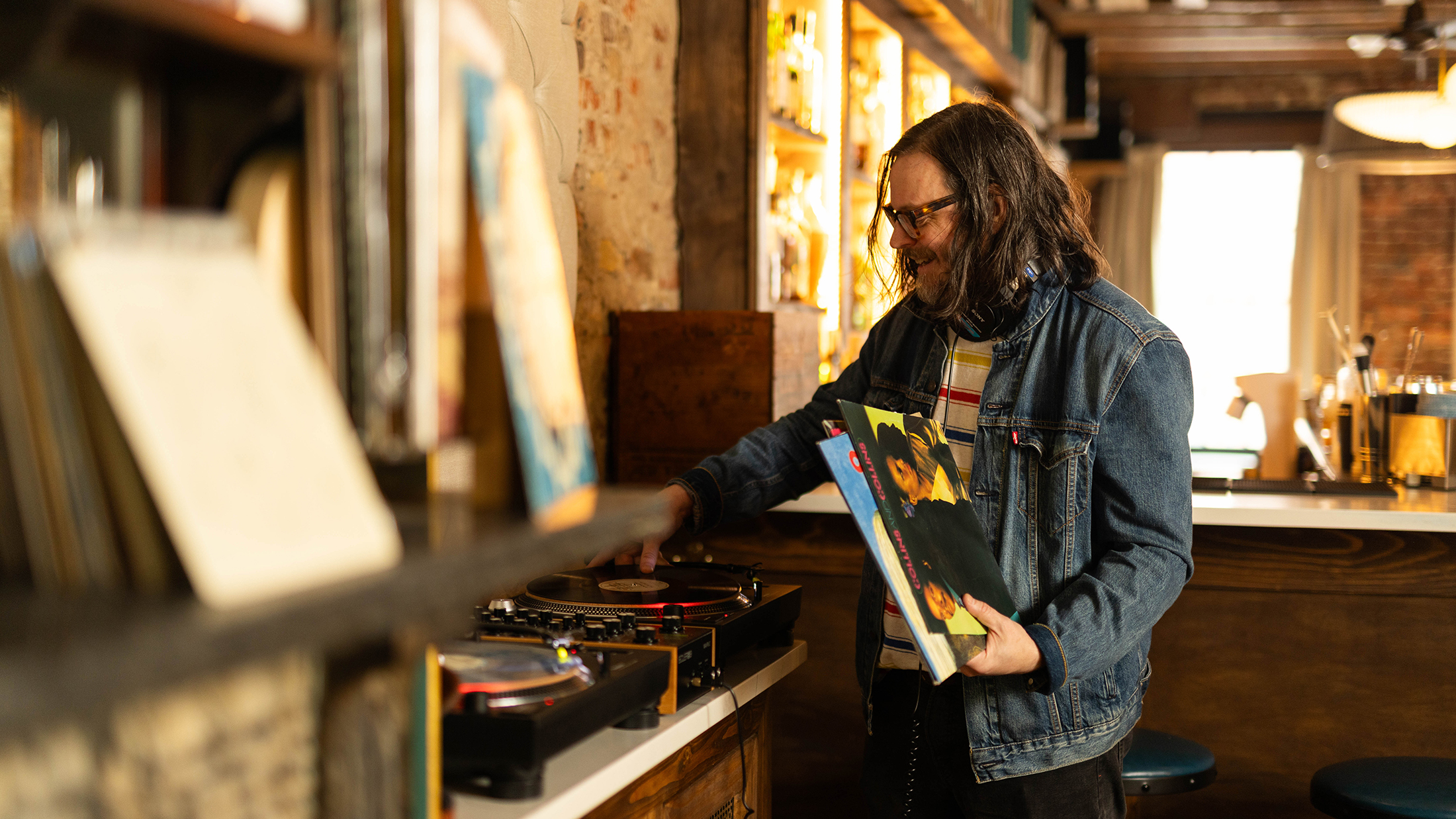 A meaning wearing a jean jacket and glasses plays a vinyl record on a turntable, while holding several record sleeves beside a shelf of vinyl records in a warmly lit bar.
