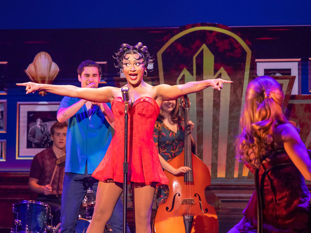 A performer in a bright red mini-dress sings into a vintage microphone with her arms outstretched, accompanied by musicians and dancers on a colorful stage.