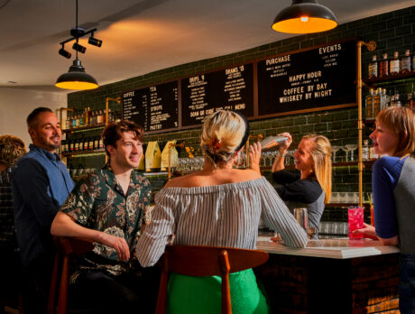 A group of guests socialize at a cocktail bar, where a bartender prepares a cocktail beneath warm pendant lighting and shelves stocked with spirits.