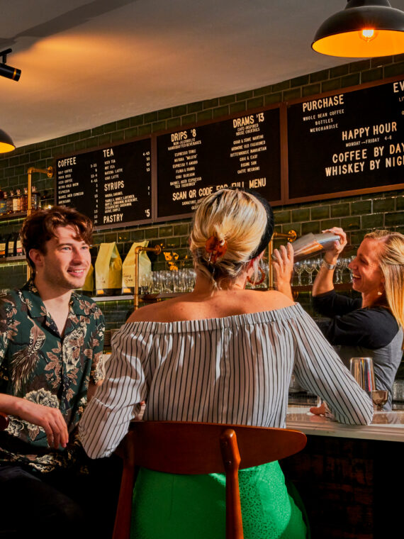 Um grupo de convidados conversa em um bar de coquetéis, onde um barman prepara um coquetel sob a luz aconchegante de luminárias pendentes e prateleiras repletas de bebidas destiladas.