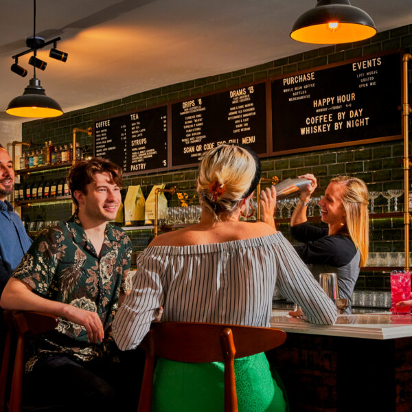 Un grupo de invitados charla en un bar de cócteles, donde un mesero prepara un cóctel bajo la cálida luz de las lámparas colgantes y entre estantes repletos de licores.