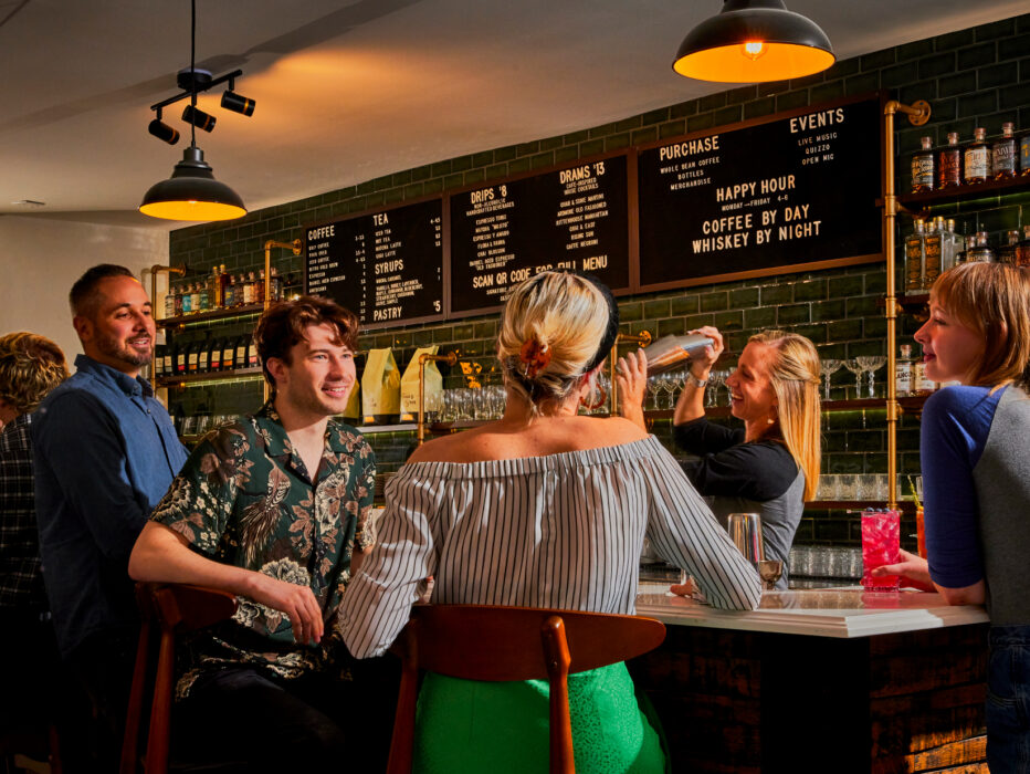 A group of guests socialize at a cocktail bar, where a bartender prepares a cocktail beneath warm pendant lighting and shelves stocked with spirits.
