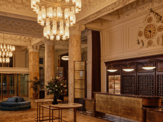 De indrukwekkende lobby van The Bellevue Hotel, met sierlijke architectonische details, marmeren zuilen, een vergulde receptiebalie en indrukwekkende kroonluchters onder een rijkelijk versierd plafond.
