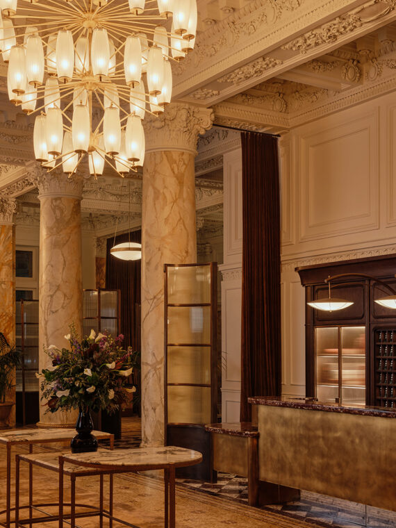 The grand lobby of The Bellevue Hotel featuring ornate architectural details, marble columns, a gilded reception desk and dramatic chandeliers beneath an intricately decorated ceiling.