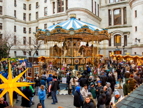 Visitors explore rows of wooden holiday market stalls decorated with glowing lights and star shaped ornaments, with a colorful Ferris wheel in the center.