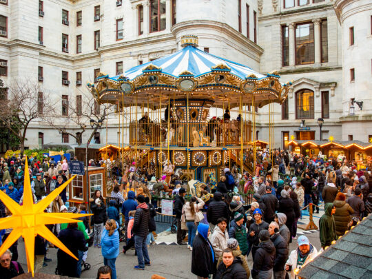 Visitors explore rows of wooden holiday market stalls decorated with glowing lights and star shaped ornaments, with a colorful Ferris wheel in the center.