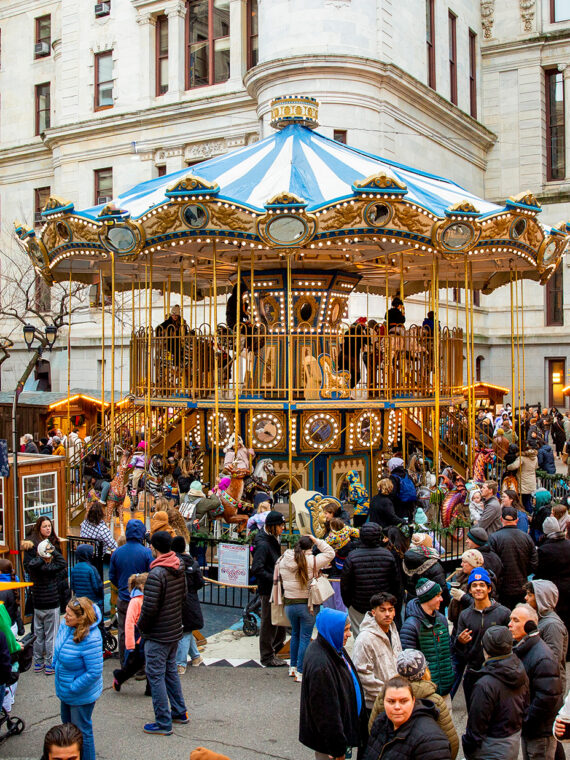 Visitors explore rows of wooden holiday market stalls decorated with glowing lights and star shaped ornaments, with a colorful Ferris wheel in the center.