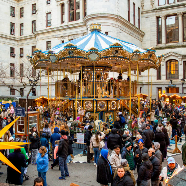 Visitors explore rows of wooden holiday market stalls decorated with glowing lights and star shaped ornaments, with a colorful Ferris wheel in the center.