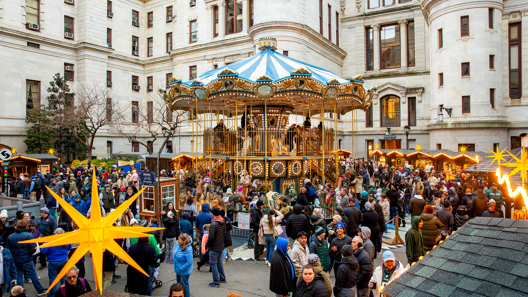 Visitors explore rows of wooden holiday market stalls decorated with glowing lights and star shaped ornaments, with a colorful Ferris wheel in the center.