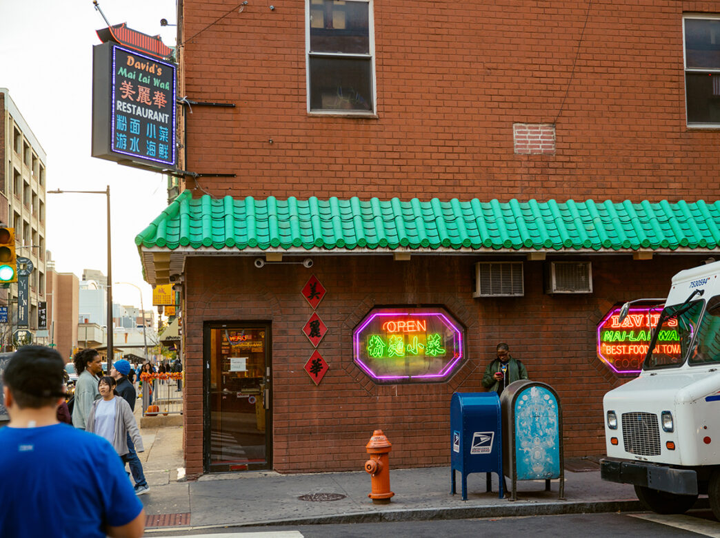 The exterior of David's Mai Lai Wah restaurant featuring a distinctive green-tiled awning, colorful neon signs and traditional Chinese lettering against a red-brick facade.