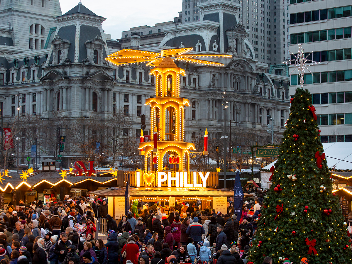 A brightly illuminated, multi-tiered German-style Christmas pyramid featuring an I Love Philly sign rises above the bustling holiday market, with a decorated Christmas tree.