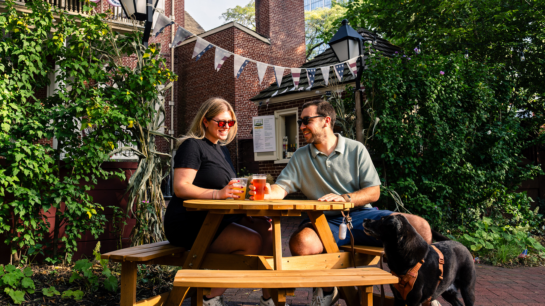 A couple enjoys drinks at a wooden picnic table with their dog in a leafy outdoor beer garden, surrounded by historic brick buildings and patriotic flags.