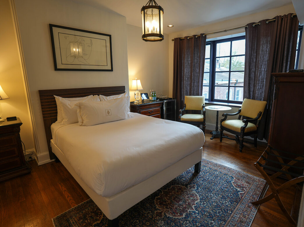 A cozy guest room featuring a queen bed with white linens, antique wood furnishings, a patterned area rug and a seating area beside a large window.