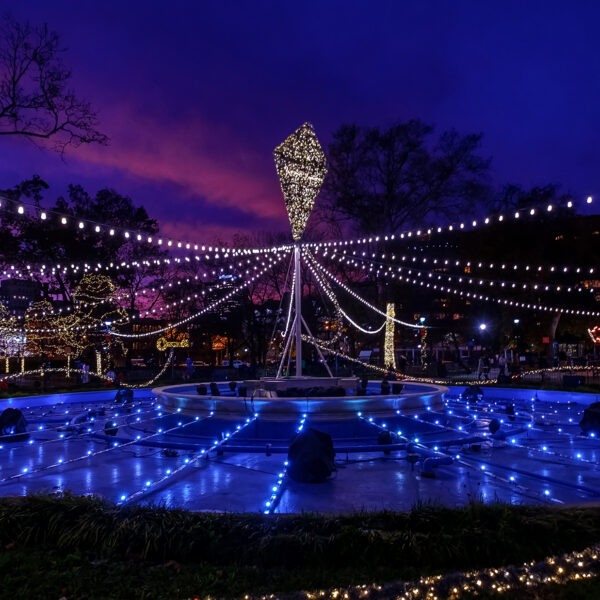Twinkling white lights stretch above an illuminated fountain at Franklin Square, with a carousel and trees shining the background beneath a purple and blue evening sky.