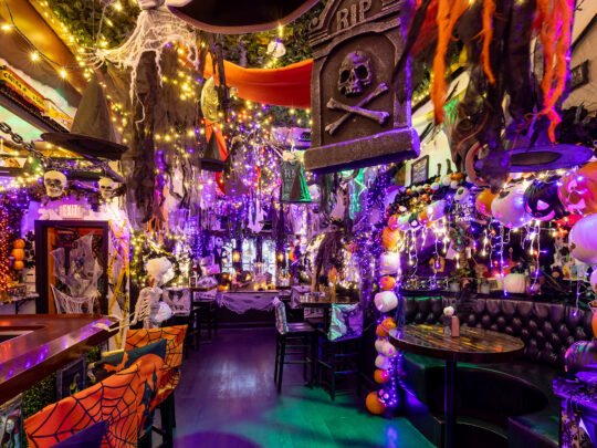 De Craftsman Row Saloon is feestelijk versierd met slingers van paarse en oranje lampjes, hangende heksenhoeden, spinnenwebben, skeletten, pompoenen en andere griezelige decoraties.