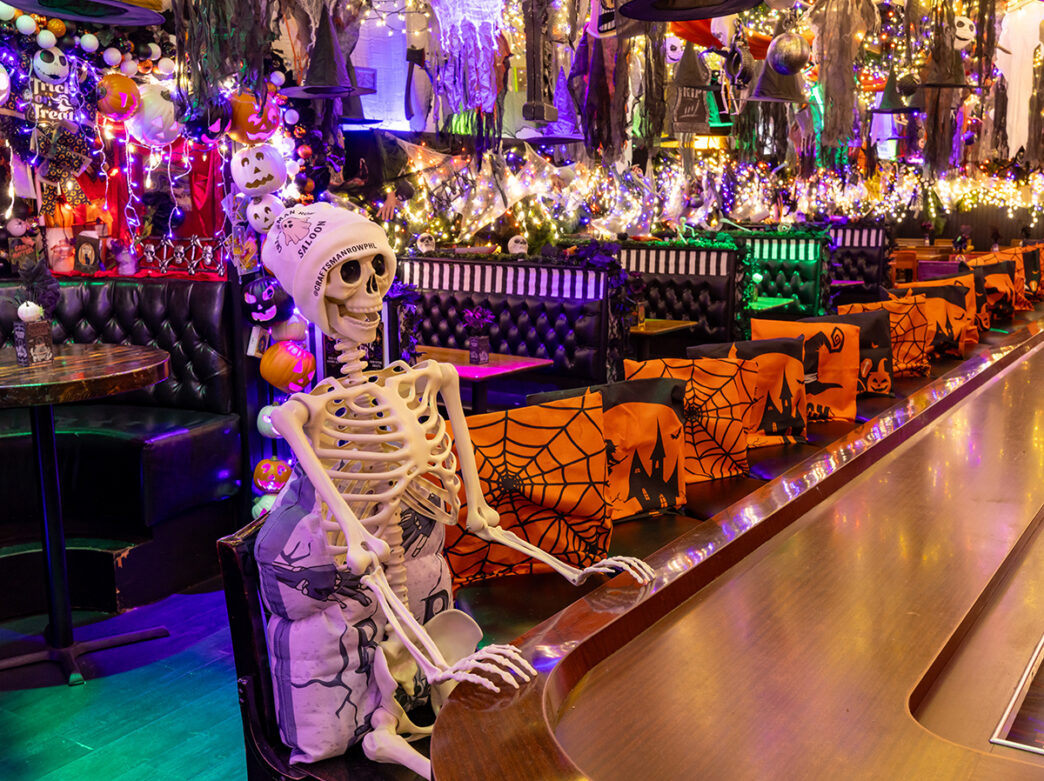 A skeleton sits at a bar inside Craftsman Row Saloon, surrounded by orange-and-black Halloween seat covers, twinkling lights, pumpkins, witches' hats and other festive decorations.