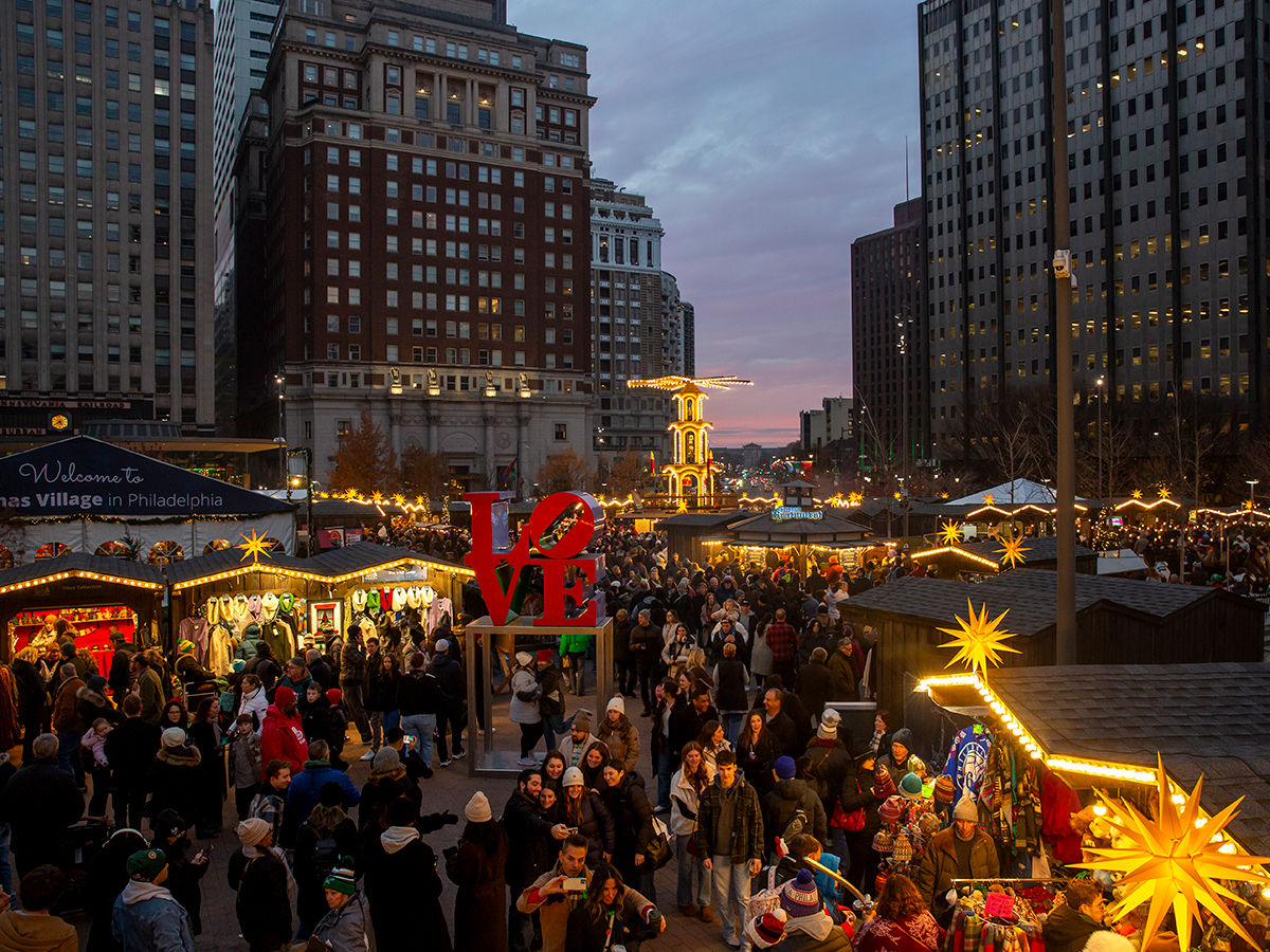 Crowds fill Christmas Village in Philadelphia at dusk, browsing illuminated wooden market stalls surrounding the iconic red LOVE sculpture and a glowing holiday pyramid.