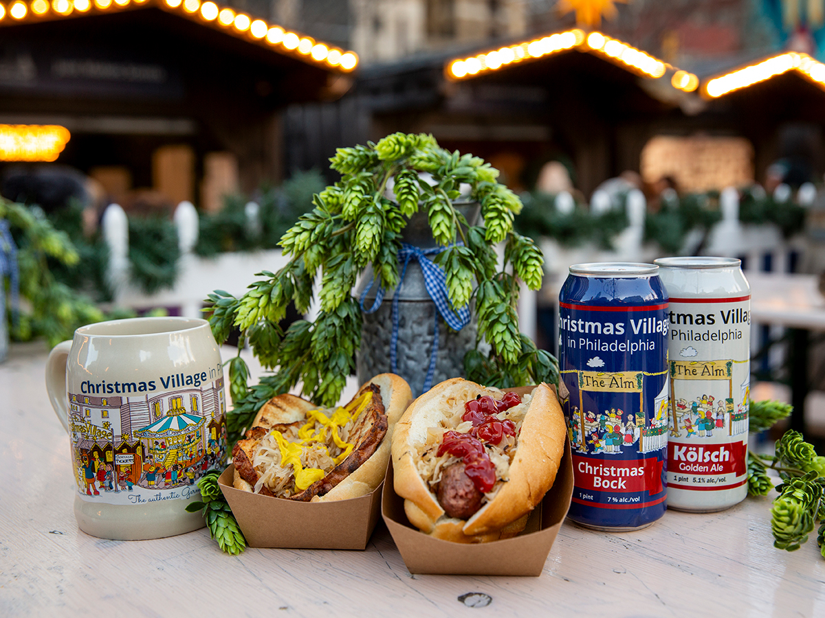 Two bratwurst sandwiches topped with sauerkraut, mustard and ketchup sit beside a Christmas Village souvenir mug and two cans of German-style beer, with festive greenery and illuminated market stalls in the background.