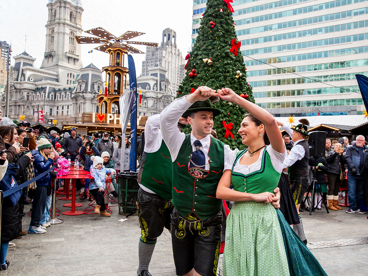 Two performers dressed in traditional German-inspired clothing dance together at Christmas Village in Philadelphia, with a decorated Christmas tree and spectators in the background.