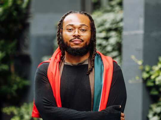 A portrait of jazz saxophonist Immanuel Wilkins wearing glasses, a black shirt and red, teal and brown silk draped over his shoulders, standing outdoors with his arms crossed in front of him.