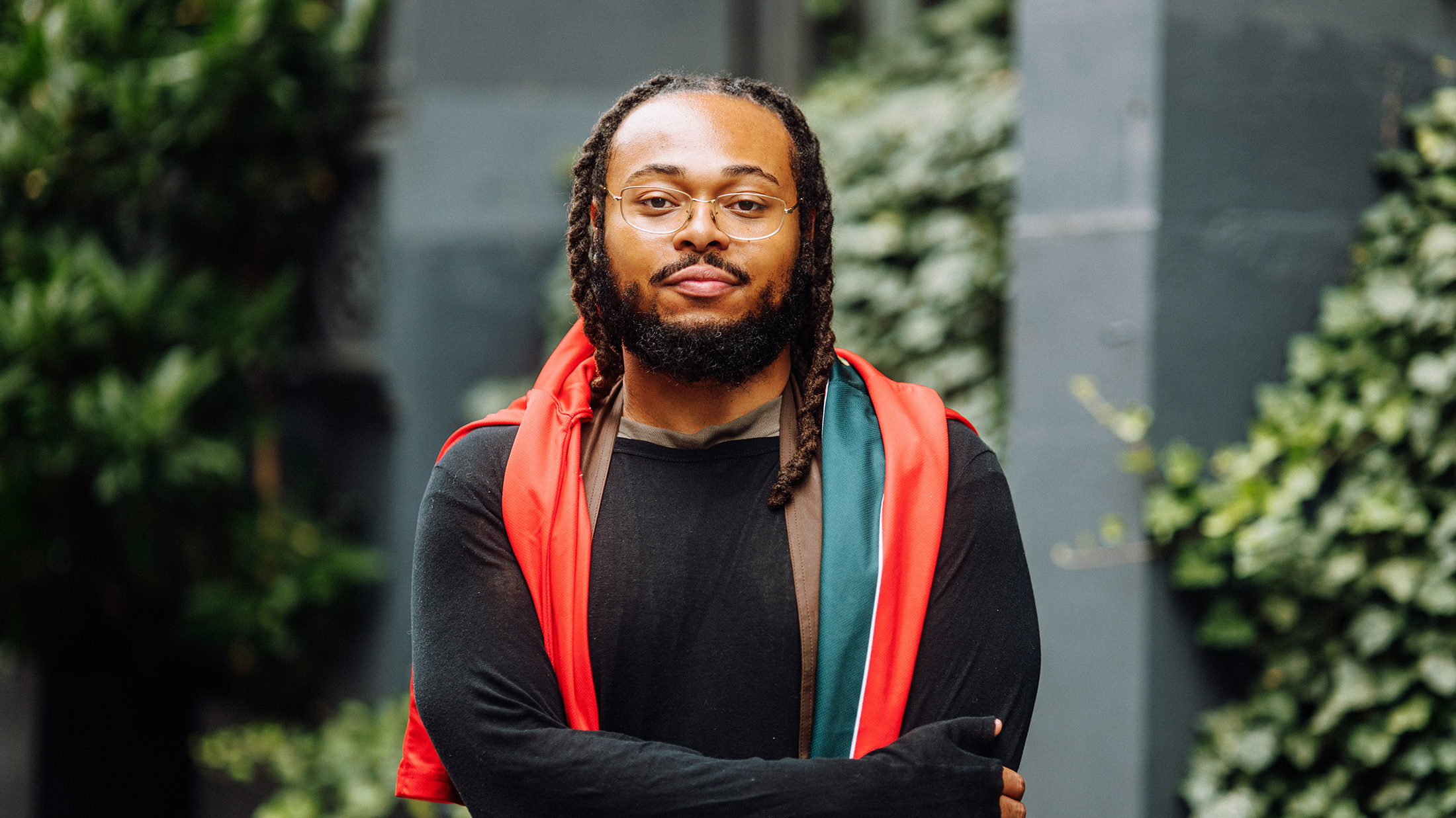 Portrait du saxophoniste de jazz Immanuel Wilkins, portant des lunettes, une chemise noire et un voile de soie rouge, bleu sarcelle et marron drapé sur les épaules, debout à l'extérieur, les bras croisés devant lui.