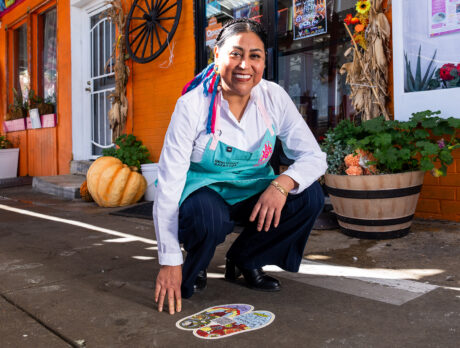 La chef Cristina Martínez lleva un delantal de color verde azulado y está agachada junto a una calcomanía de una huella de pie de colores en la acera, frente a un local de color naranja brillante.