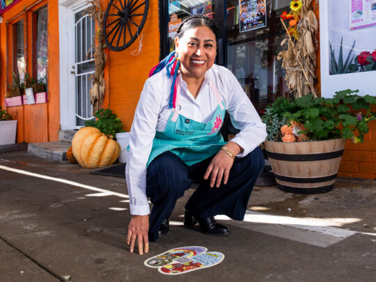 La chef Cristina Martínez lleva un delantal de color verde azulado y está agachada junto a una calcomanía de una huella de pie de colores en la acera, frente a un local de color naranja brillante.