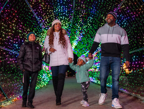 A family wearing winter jackets and beanies walks hand-in-hand through a tunnel of colorful holiday lights at night.