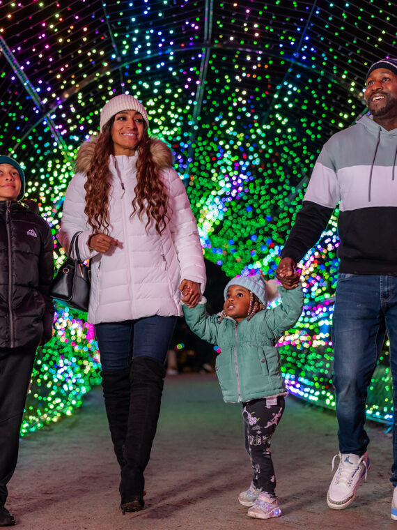 Una familia ataviada con chaquetas y gorros de invierno camina de la mano por un túnel de luces navideñas de colores por la noche.