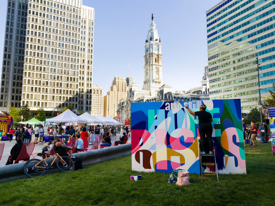 An artist paints a colorful mural while standing on a step ladder in a grassy lawn, with vendor tents and Philadelphia's City Hall in the background.