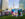 An artist paints a colorful mural while standing on a step ladder in a grassy lawn, with vendor tents and Philadelphia's City Hall in the background.