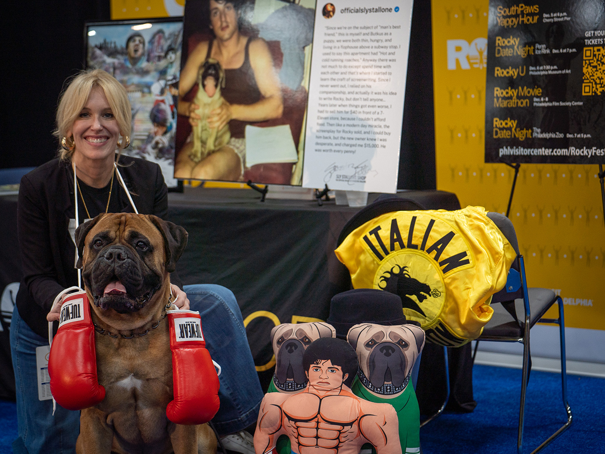 A large bullmastiff with red boxing gloves hanging from their neck sits with a woman beside Rocky-themed props, including a yellow "Italian" robe, black hat and illustrated character pillows.