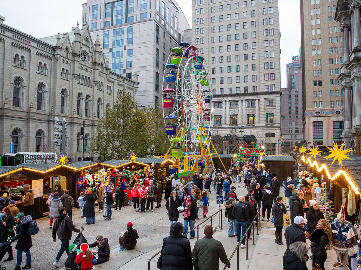 A colorful Ferris wheel rises above busting holiday market stalls, where visitors browse vendors beneath illuminated star decorations and surrounding Center City Buildings.