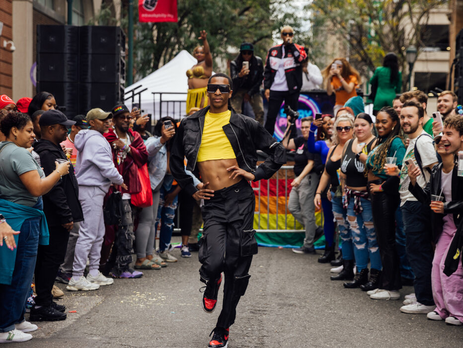 A person wearing a yellow crop top, black jacket, black pants and sunglasses struts through the street while a crowd cheers on during OurFest in Philadelphia.