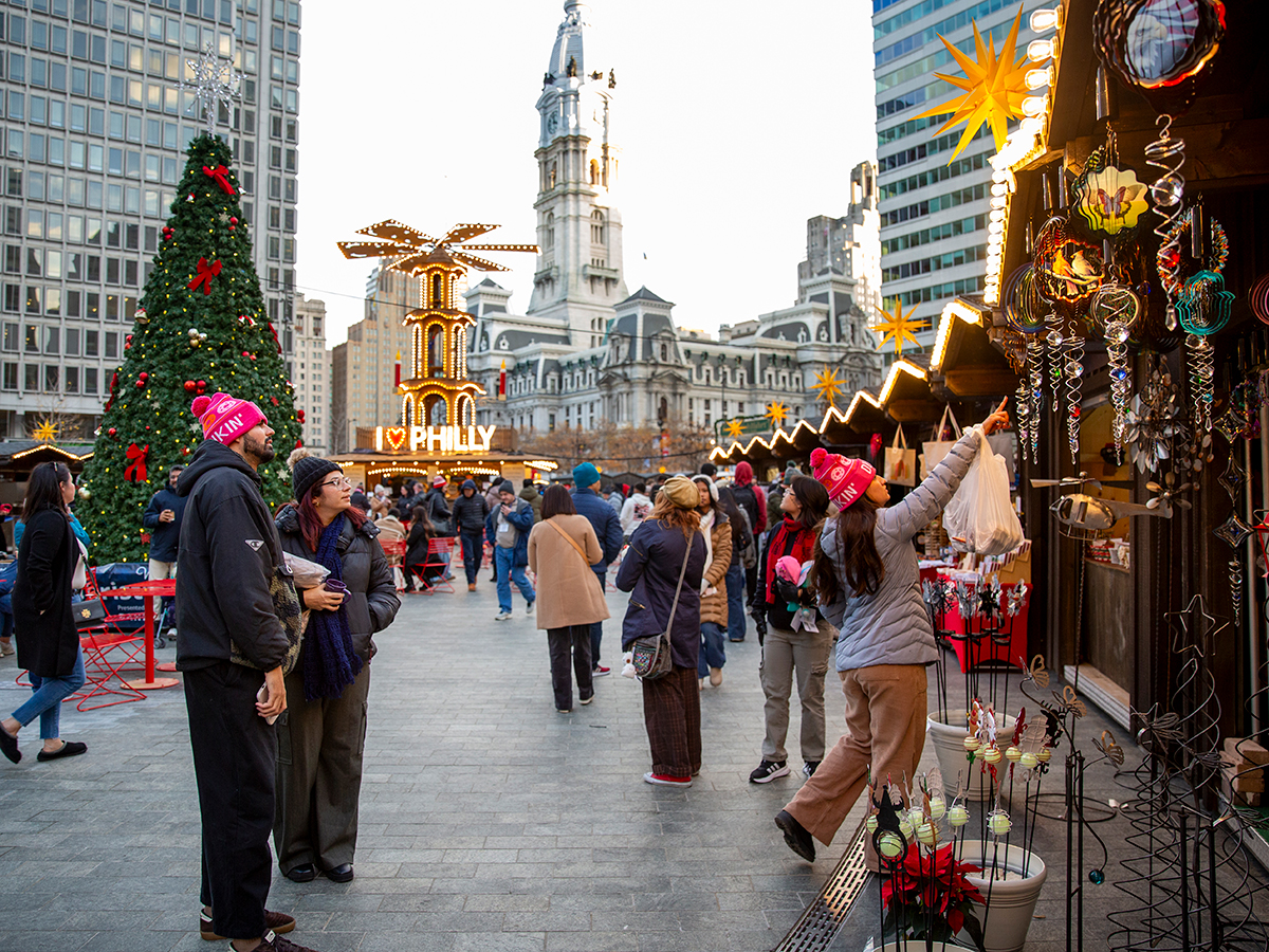 Shoppers browse colorful hanging ornaments and handcrafted decorations at a wooden market stall, with a towering Christmas tree, illuminated holiday pyramid and City Hall in the background.