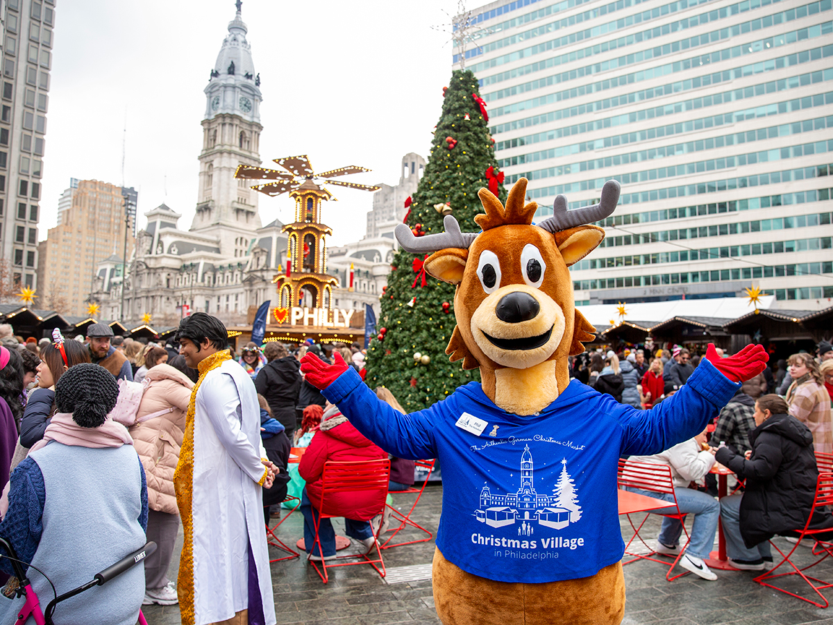 A reindeer mascot spreads his arms in front of a towering Christmas tree, with the illuminated holiday pyramid and City Hall in the background, as crowds fill the Christmas Village.