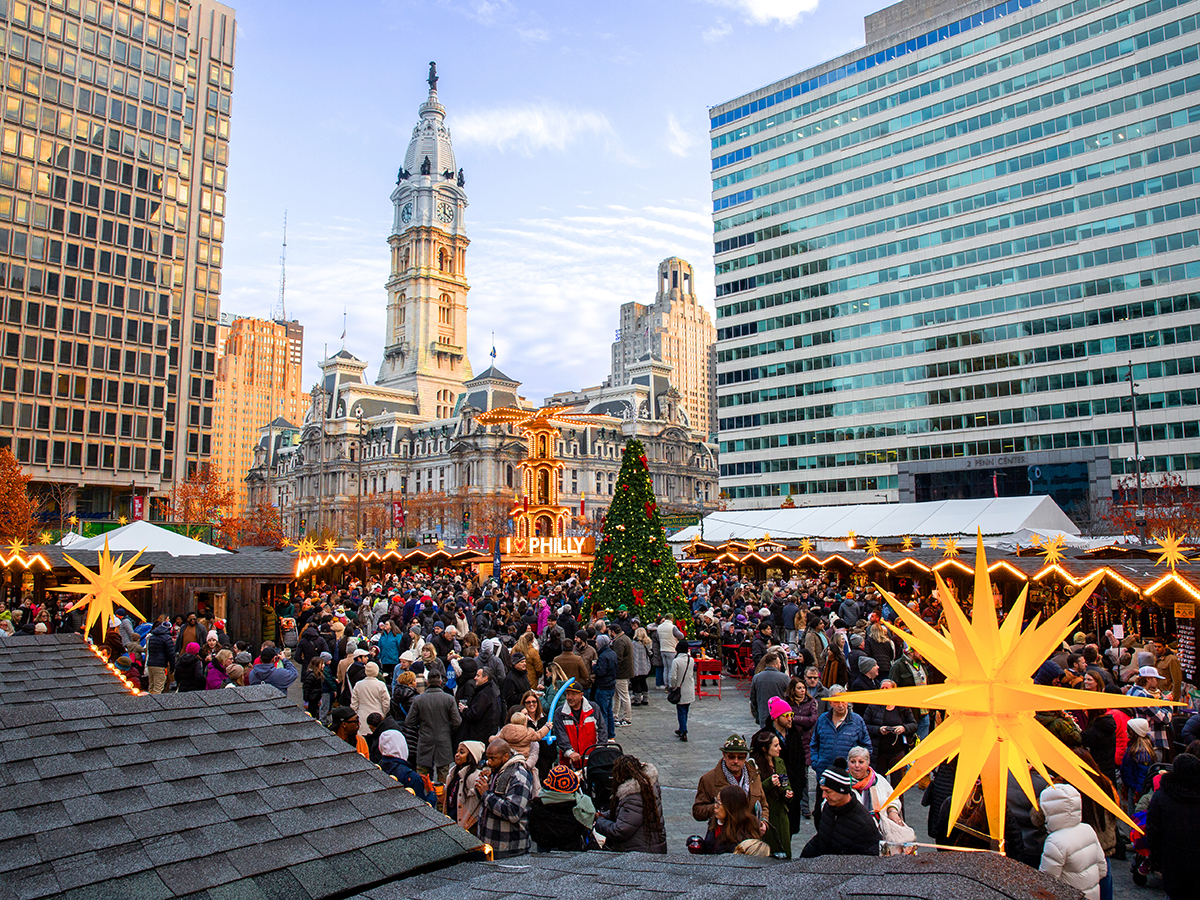 Vendor huts, a large Christmas tree and glowing German-style holiday pyramid are set up in Love Park for Philadelphia's Christmas Village. People walk around shopping and taking photos. City Hall is in the background.