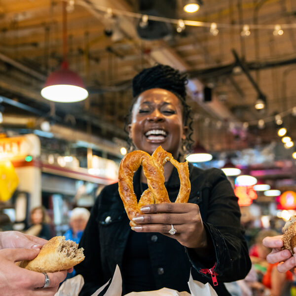 Drie bezoekers lachen gezellig samen terwijl ze genieten van populaire gerechten uit Philadelphia, waaronder een versgebakken zachte pretzel, een broodje geroosterd varkensvlees en een cheesesteak bij Reading Terminal Market.